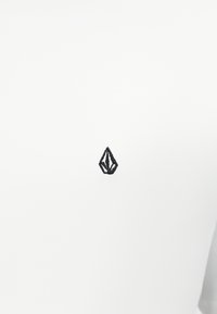 Άσπρο μπλουζάκι με μαύρο κεντημένο λογότυπο σε σχήμα μυτερού διαμαντιού. Το ύφασμα φαίνεται λείο και ελαφρύ.