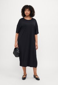 Maksi gaun hitam yang terbuat dari kain lembut, menampilkan leher bulat, lengan pendek, dan potongan santai, dipadukan dengan tas hitam bertekstur.