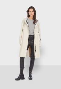 Světle béžový plášť s kapucí, na knoflíky, se stahovacími šňůrkami, v kombinaci se šedou košilí a černými kalhotami z umělé kůže. Černé kotníkové boty.