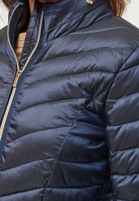 Navy blue puffer jacket made from glossy material with quilted pattern, full zip closure, and gold-tone hardware at the collar.