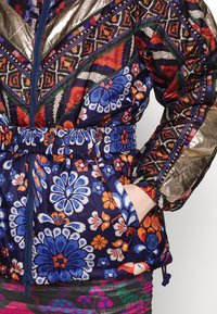 Veste avec des motifs floraux et géométriques colorés en bleu, orange, rouge et or, dotée d'une fermeture éclair avant et d'une taille élastique, main dans la poche.