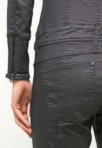 Gros plan d'une personne portant un pantalon en cuir noir et une veste assortie avec un poignet zippé, main reposant sur le côté.