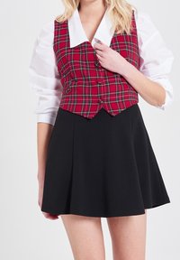 Czerwono-zielona kamizelka w kratę z białym kołnierzykiem i guzikami, zestawiona z czarną, rozszerzaną spódnicą typu skater. Gładki materiał bez widocznych wzorów.