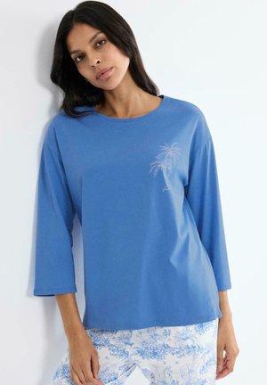 Frau trägt ein blaues Langarmshirt mit kleinem Palmenmuster und weiße Hosen mit blauen Blumenmustern, steht vor einer weißen Wand.
