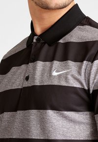 Polo rayé noir et gris avec un tissu texturé, doté d'une patte de boutonnage et d'un col noir. Logo Nike sur le côté gauche.