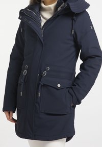 Chaqueta exterior de color azul marino en un tejido suave y texturizado. Cuenta con capucha, múltiples bolsillos con cierres de tipo toggle y acentos de hardware plateado.