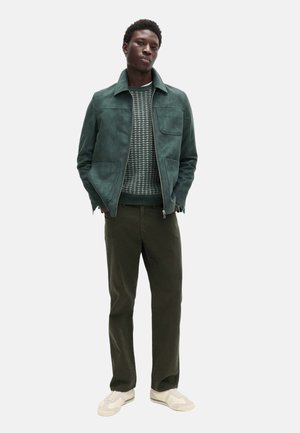 Giacca in camoscio verde con zip frontale, maglione lavorato a maglia testurizzato, pantaloni oliva e sneakers beige. Modello in piedi con le mani nelle tasche.