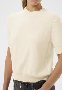 Kremeni pulover s kratkim rukavima, s rebrastim izrezom i rubom. Pletena tekstura, malo opušteni kroj. Nositi s crnim pojasom i trapericama.