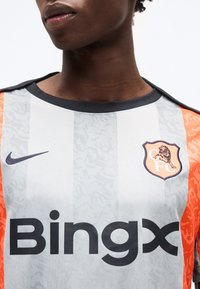 Κοντινό ενός ποδοσφαιρικού jersey με λευκές και γκρι ρίγες, πορτοκαλί μανίκια, μαύρο λογότυπο Nike, χορηγό BingX και έμβλημα λιονταριού με τα γράμματα "CFC".