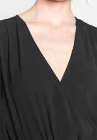 Svart omlottklänning med djup V-urringning, mjuk textur och lätta veck på axlarna. Tyget är lätt och flödar.