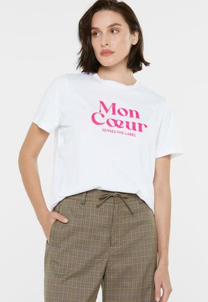 White cotton t-shirt featuring bold pink "Mon Coeur" text. Worn with patterned beige and brown check trousers, displaying a relaxed fit.