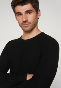 Schwarzes, geripptes Langarmshirt mit rundem Ausschnitt. Das Modell posiert mit verschränkten Armen und zeigt dabei die Passform und die Textur des Stoffs.