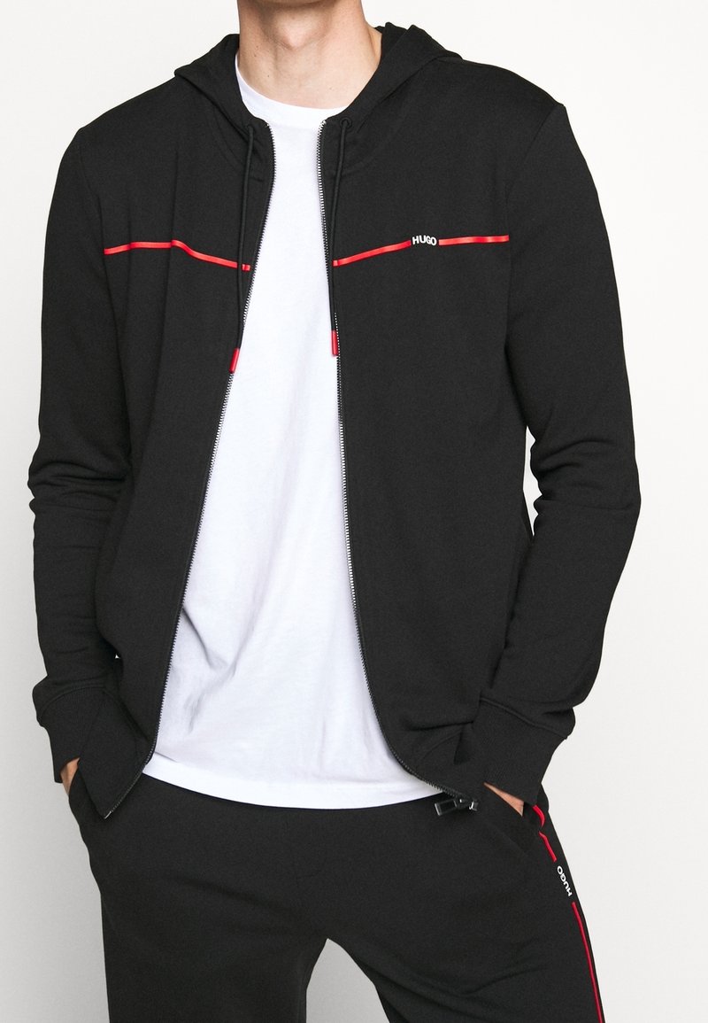 Uomo che indossa una felpa nera con zip e pantaloni neri coordinate con accenti rossi, oltre a una maglietta bianca sotto, con le mani nelle tasche.