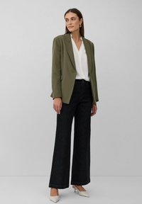 Blazer vert olive sur une blouse blanche, associé à un pantalon en denim foncé à jambes larges et des talons crémeux, mettant en valeur un design ajusté et des lignes épurées.