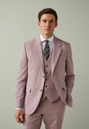 Man wearing a light pink three-piece suit with a white shirt and black striped tie, standing against a plain light background.