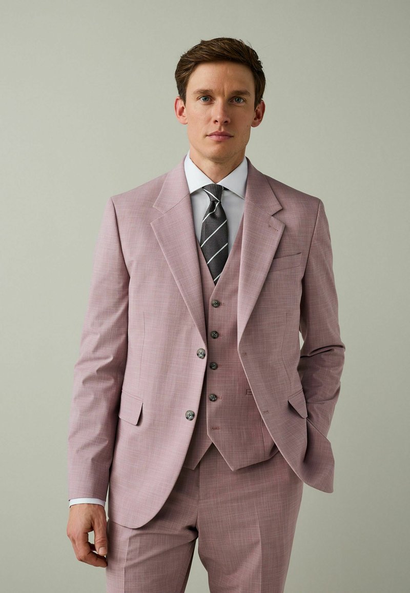 Egy férfi világos rózsaszín háromrészes öltönyt visel, fehér inggel és fekete csíkos nyakkendővel, egyszínű világos háttér előtt áll.