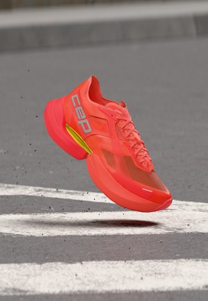 Bright orange running shoe with yellow accent, floating above asphalt with white road markings and scattered small gravel.