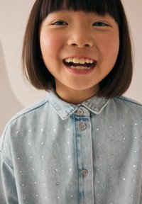 Camicia in denim azzurro chiaro con colletto e dettagli in metallo argentato, indossata da un bambino sorridente.