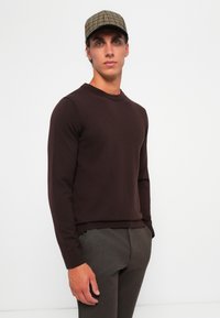 Man wearing a brown sweater, gray pants, and a checkered cap, standing with one hand in pocket against a plain white background.