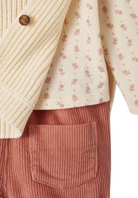 Gestrickte cremfarbene Strickjacke mit geripptem Muster und einem Holzknopf, über einem hellbeigen Hemd mit pinkem Blumenmuster und rostfarbenen Cordhosen mit Tasche.