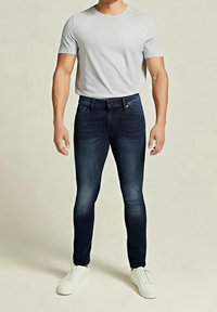 Jeans ajustados de mezclilla oscura con un ligero desvanecido, combinados con una camiseta de manga corta gris claro y zapatillas blancas. Diseño simple, sin adornos.