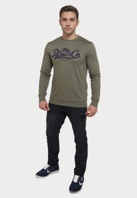 Sudadera de manga larga verde oliva con texto y logotipo en azul marino, combinada con jeans oscuros y zapatillas azul marino. Diseño simple y ajustado con puños acanalados.