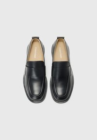 Crne kožne loafers cipele s širokom trakom sprijeda, zaobljenim vrhom i suptilnim detaljima šivanja. Udobna unutrašnjost, minimalistički dizajn.