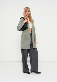 Világosszürke kötött kardigán elülső zsebekkel, szürke, bő szárú nadrággal és fekete, hegyes orrú cipővel párosítva. Egy fekete táska van a vállon átvetve.