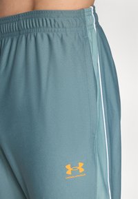 Lyseblå treningbukser med teksturert stoff, elastisk livbånd, hvite sidelister og en oransje Under Armour-logo på låret.