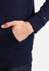 Marinblå sweatshirt med kenguruficka, ribbade ärmslut och nederkant. Har en liten röd, vit och blå logotyp på ärmen.