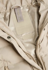 Beżowa kurtka puchowa od wewnątrz z metalową siateczkową podszewką, metka wskazująca rozmiar "SMAŁY" oraz zamek błyskawiczny z metalowymi elementami.