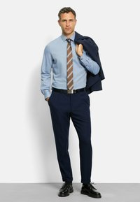 Herrenoutfit bestehend aus einem hellblauen Hemd, einer braun gestreiften Krawatte, marineblauen Hosen und schwarzen Schuhen; komplettiert mit einem marineblauen Blazer, der über dem Arm getragen wird.