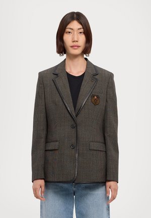 Giovane che indossa un blazer marrone a quadri con rifiniture nere, uno stemma sulla tasca del petto, una camicia nera e jeans azzurri su uno sfondo semplice.