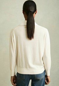 Helles beige Strickpullover mit Kragen, langen Ärmeln und gerippten Bündchen. Kombiniert mit dunkelblauen Jeans im lässigen Schnitt.