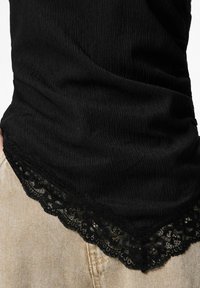 Melns tops ar teksturētu audumu un mežģīņu apdari apakšmalā, radot slāņotu efektu virs gaiši bēšām biksēm.