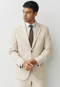 Lichtbeige kostuum gemaakt van een getextureerde stof, met een enkele knoop, notch-lapels en een borstzak; gecombineerd met een wit shirt en een bruine stropdas.