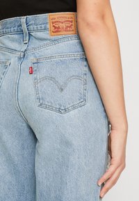 Světlé modré džíny z denimu s koženou značkovou nášivkou, charakteristickými zadními kapsami a kontrastním prošíváním. Všimněte si detailu postranního švu.