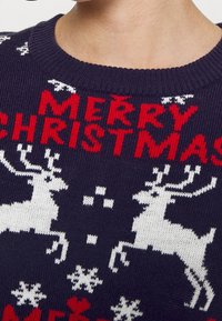 Marinblå stickad tröja med röd "GOD JUL"-text, vita renar och snöflingor i ett festligt mönster. Rund halsringning och elastiska ärmslut.