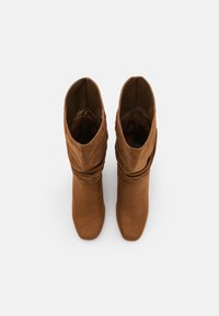 Stivaletti in suede marrone con un design morbido, punta affusolata e texture liscia. Presentano un'altezza fino al polpaccio e pieghe morbide e casual.