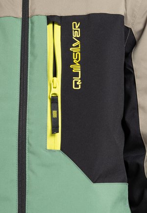 Hybrid-Jacke mit grünen, schwarzen und beige Panels, ausgestattet mit einem auffälligen gelben Reißverschluss und dem "Quiksilver"-Logo in leuchtendem Gelb auf der Vorderseite.