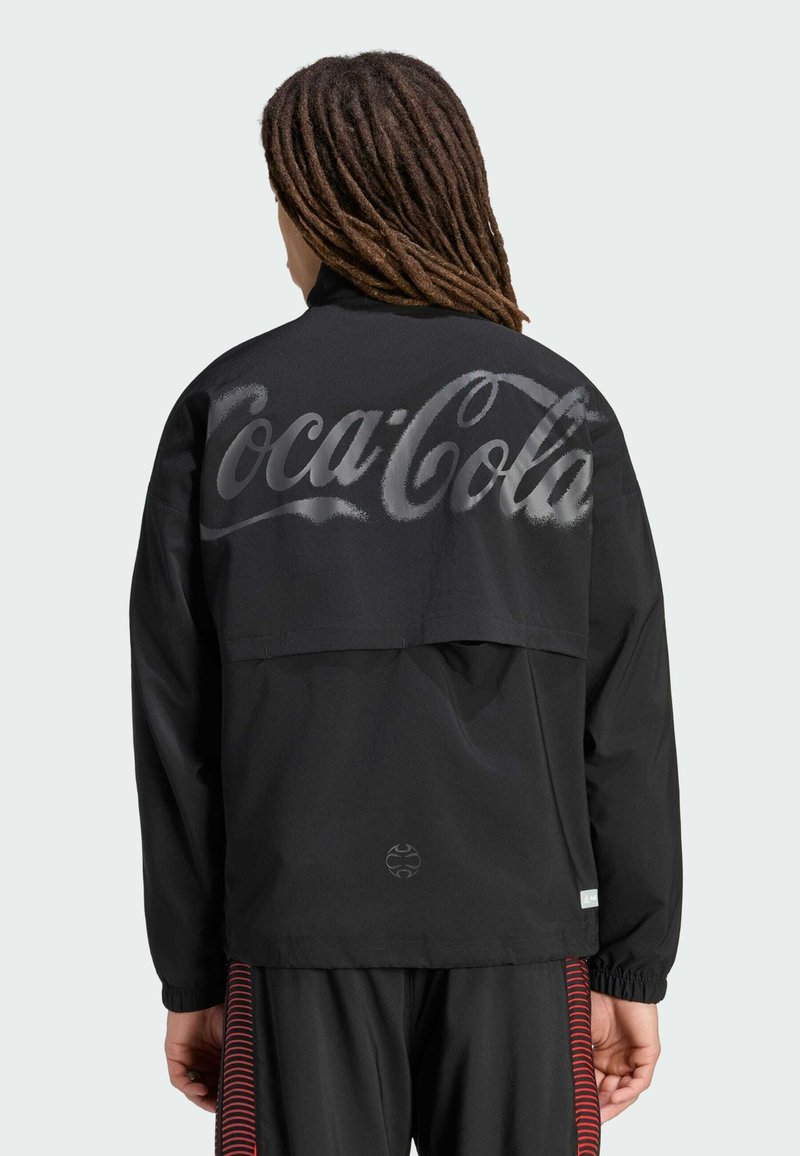 Person with long dreadlocks wearing a black jacket featuring large, faded "Coca-Cola" text across the back and black pants with red stripes.