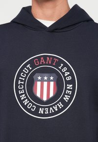 Tengerészkék kapucnis pulóver kör alakú grafikával, amelyen pajzs, piros és fehér csíkok, valamint a "GANT 1949 NEW HAVEN CONNECTICUT" felirat található.