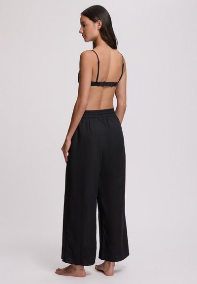 Γυναίκα που στέκεται ξυπόλυτη, βλέποντας πλάγια, φορώντας μαύρο σουτιέν και άνετο μαύρο παντελόνι με φαρδιές ίσιες γραμμές και ψηλή μέση, σε απλό φόντο.