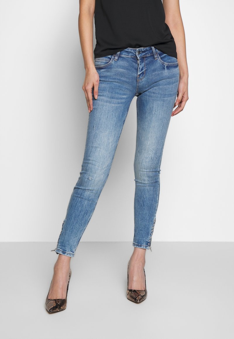 Jean skinny bleu avec un délavage usé, bords de chevilles effilochés et boutons décoratifs sur les côtés, associé à un top noir et des talons en imprimé serpent.