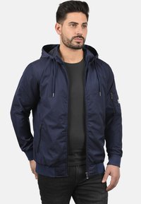 Mørkeblå hettejakke laget av lett stoff, med frontglidelås, sidelommer og elastiske mansjetter, designet med et minimalistisk estetikk.