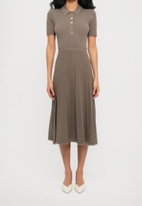 Femme portant une robe en maille côtelée taupe avec manches courtes, un col polo boutonné et une jupe midi plissée, associée à des chaussures blanches pointues.