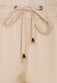 Detailní záběr na béžové látkové kalhoty s elastickým pasem a zavázanou šňůrkou s kovovými koncovkami.