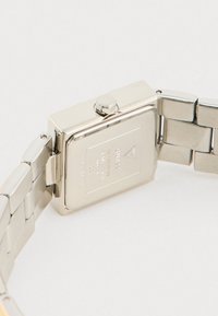 Ceas de mână din metal argintiu, cu un cadru rectangular, coroana plată și o brățară cu zale. Logo-ul mărcii gravat pe spatele carcasei.
