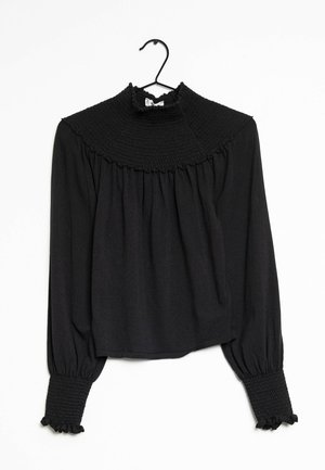 Blusa negra de manga larga con cuello alto fruncido y puños, colgada en una percha negra sobre un fondo blanco.