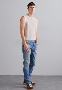 Svetlo bež pletena brezrokavnik v kombinaciji s svetlo modrimi jeans hlačami, ki imajo sproščen kroj, obut v rjave superge z belimi vezalkami.